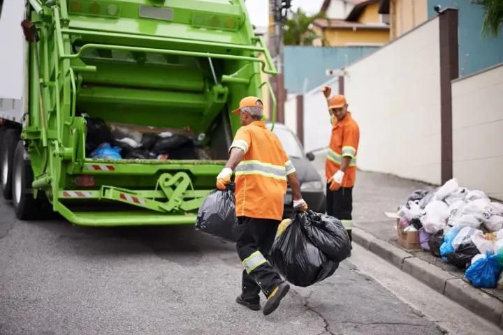 Garbage collectors in orange vests load black trash bags into a green sanitation truck on a street