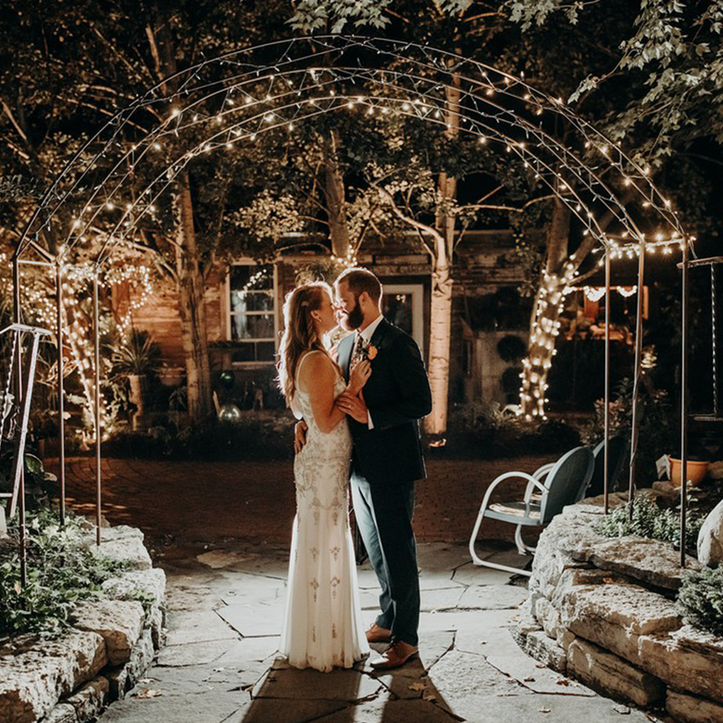 A bride and groom are kissing under an archway with lights.