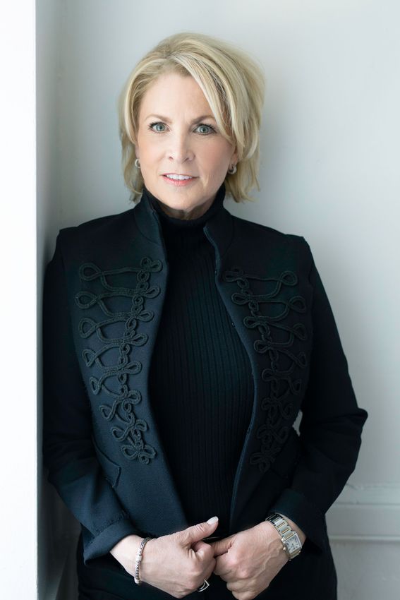 Blonde woman in black outfit, leaning against a white wall, smiling.