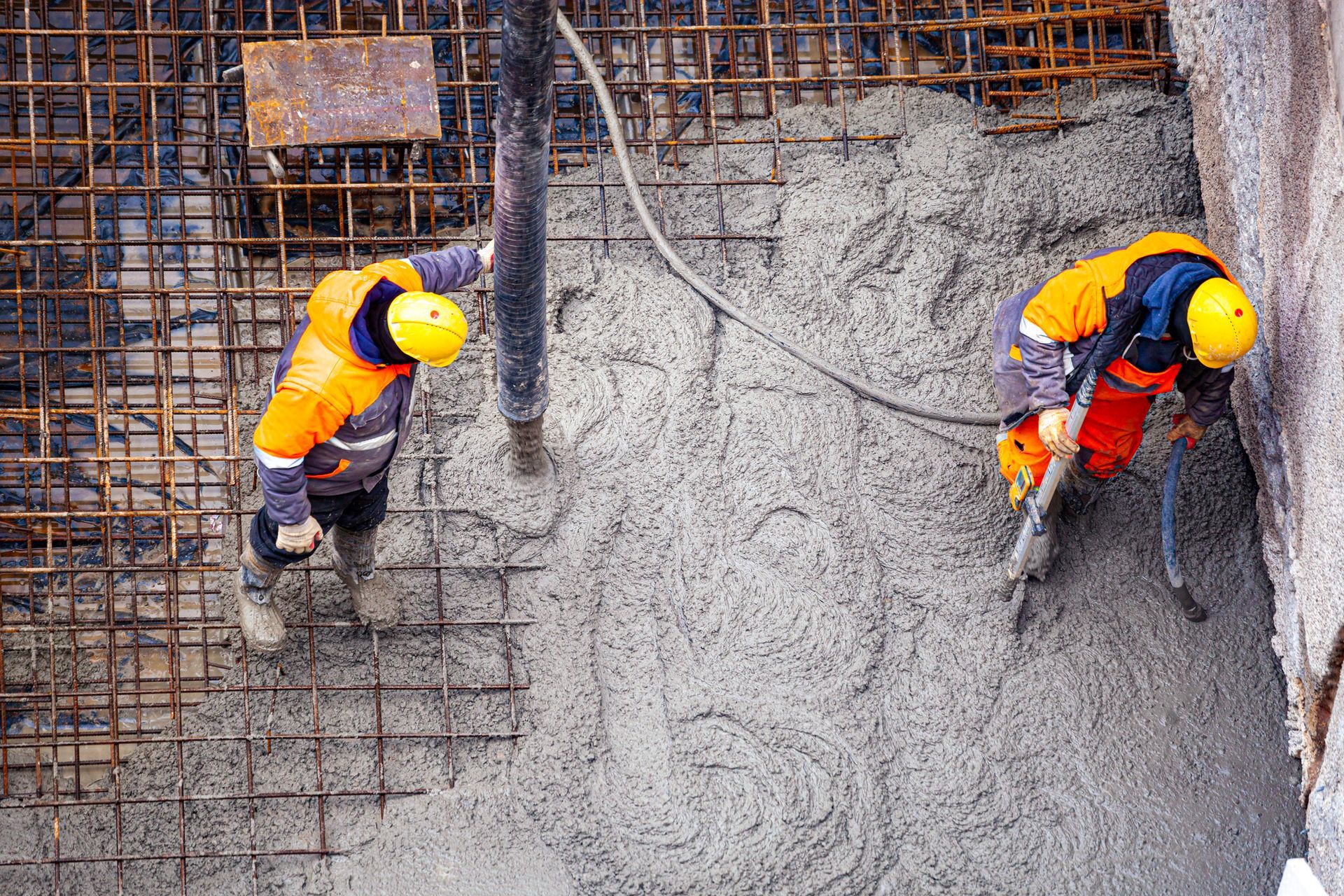 Two workers in safety gear are pouring cement on a rebar grid floor during concrete construction.