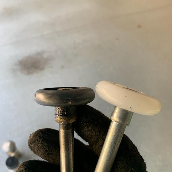 Two garage door rollers: one worn black, one new white, held by gloved hand.