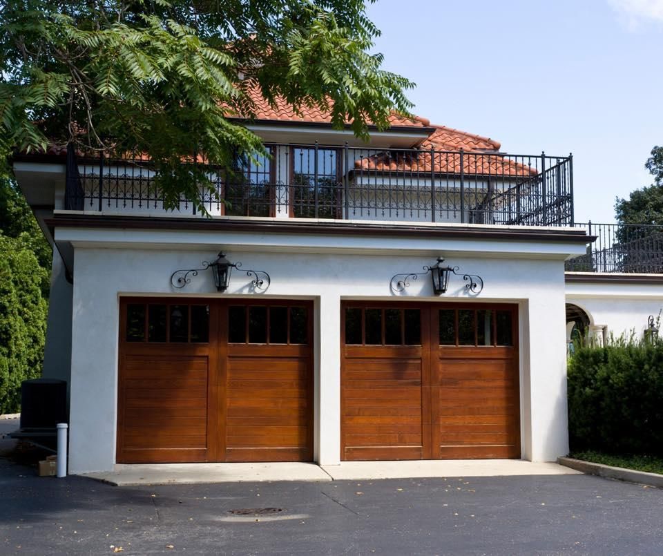 Two-car garage with brown doors, a white exterior, and a balcony with a wrought-iron railing.