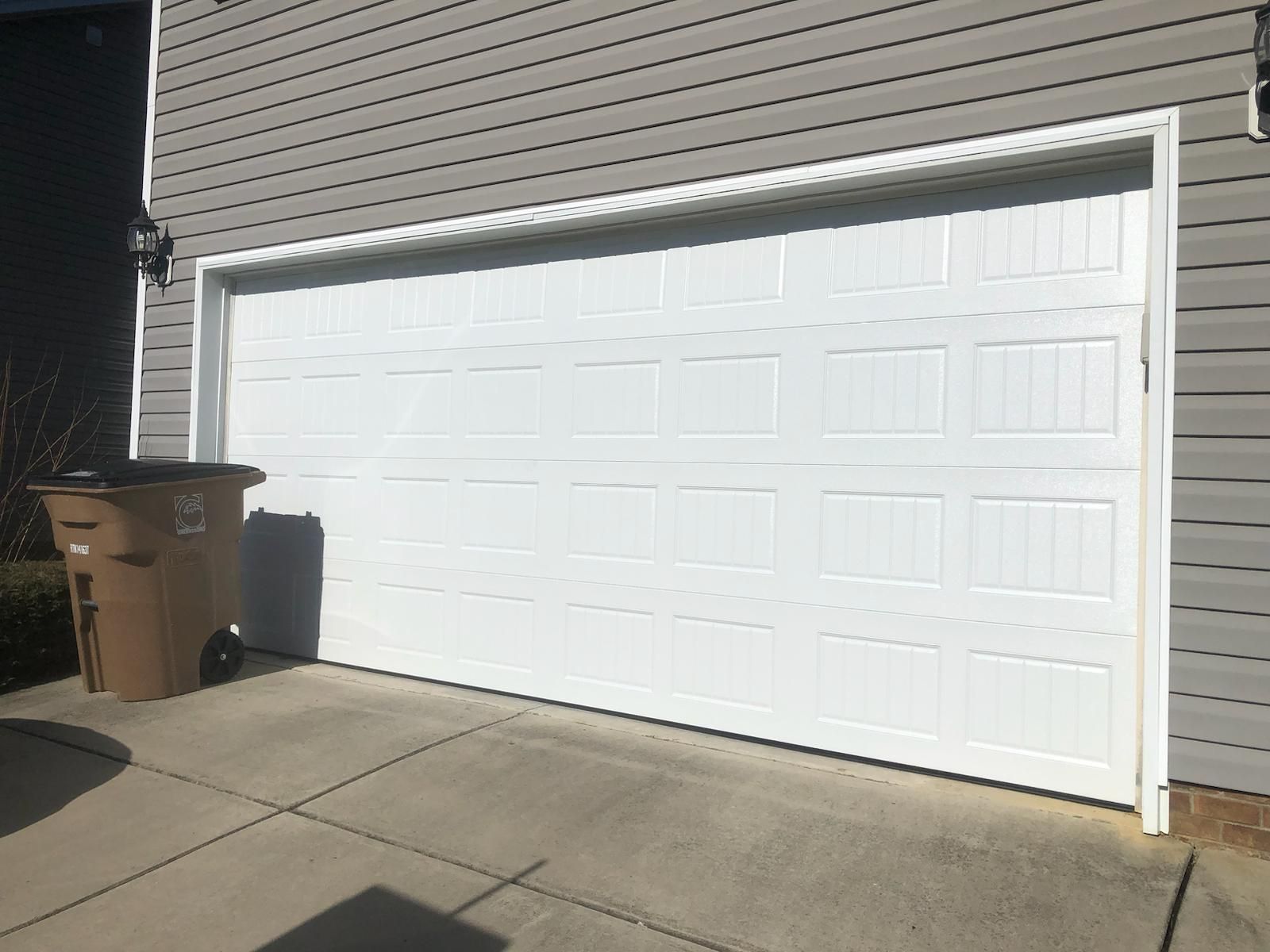 White garage door on a gray house, brown trash can in front.