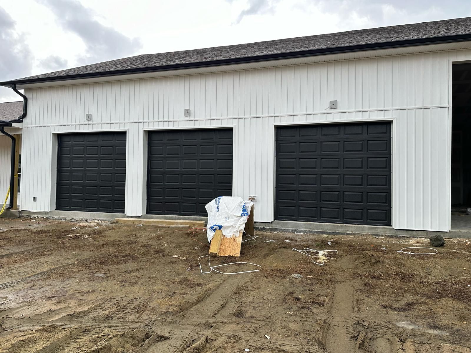 Three-bay garage with black doors and white siding on a muddy construction site.