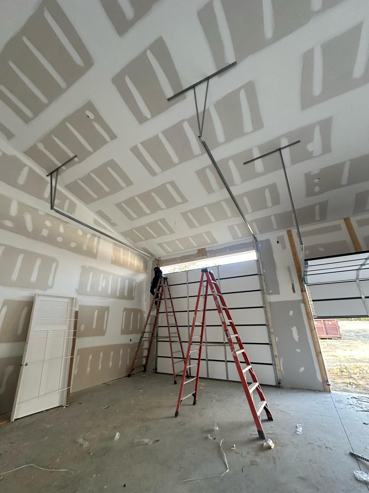 Garage interior under construction: drywall installed on walls and ceiling, two ladders, person working.