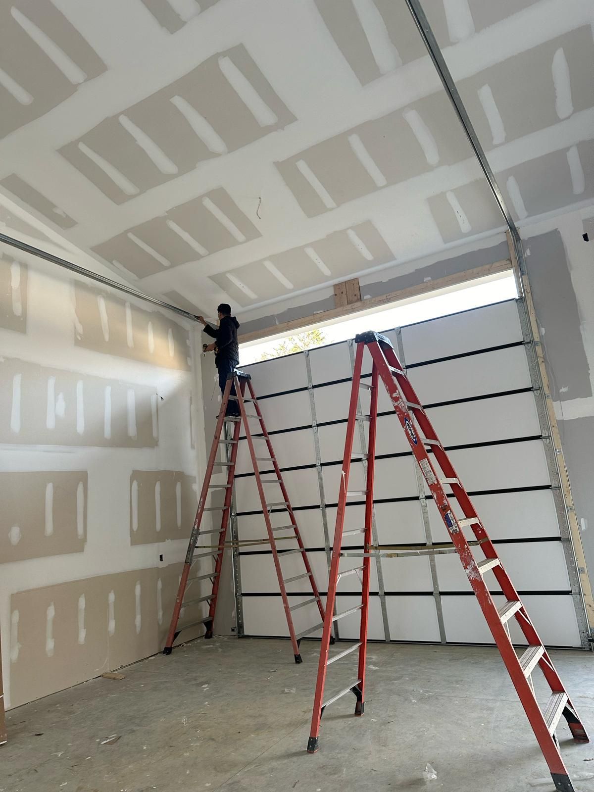 Person on a ladder installing trim in a garage under construction; drywall and garage door visible.