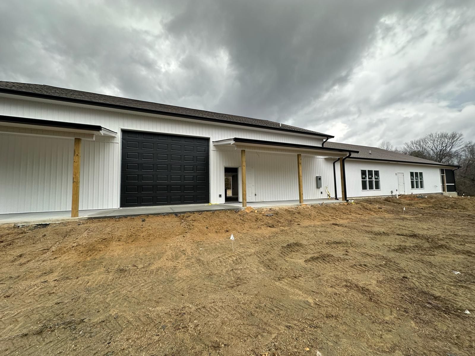 New white building with black garage door, brown roof, and wooden supports on a cloudy day.
