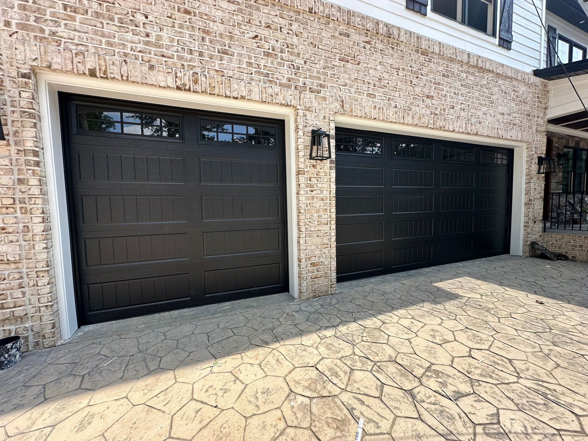 Two black garage doors with windows, on a house with brick exterior and stone driveway.