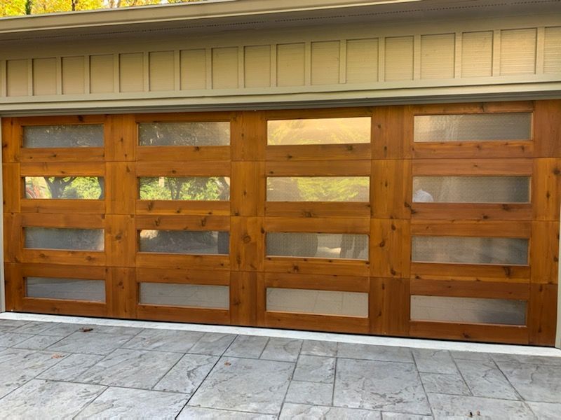Wooden garage door with rectangular glass panels, set in a light-colored building, over a concrete driveway.