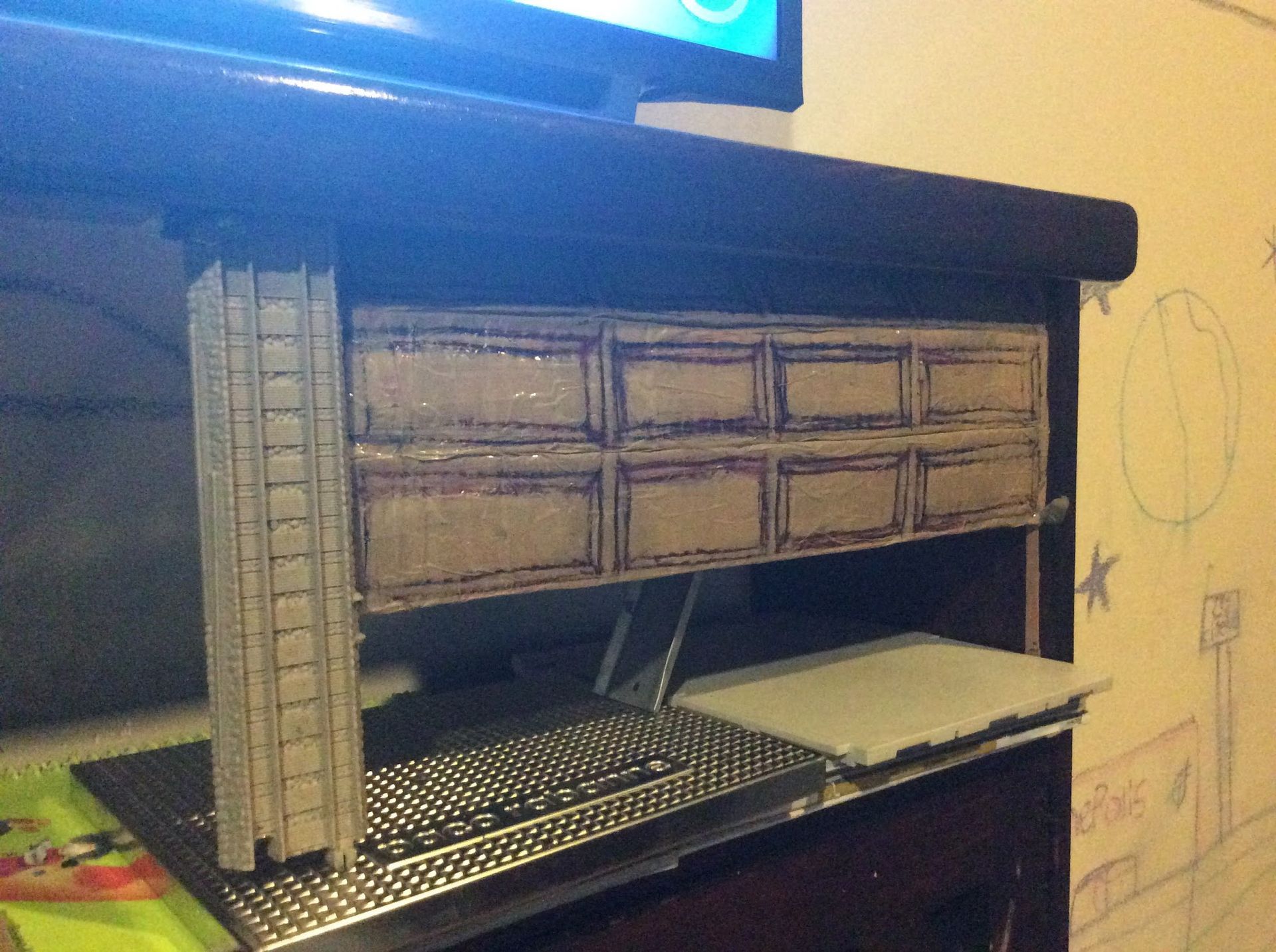 Model skyscraper next to a building facade on a tabletop, near a screen and a drawing on the wall.