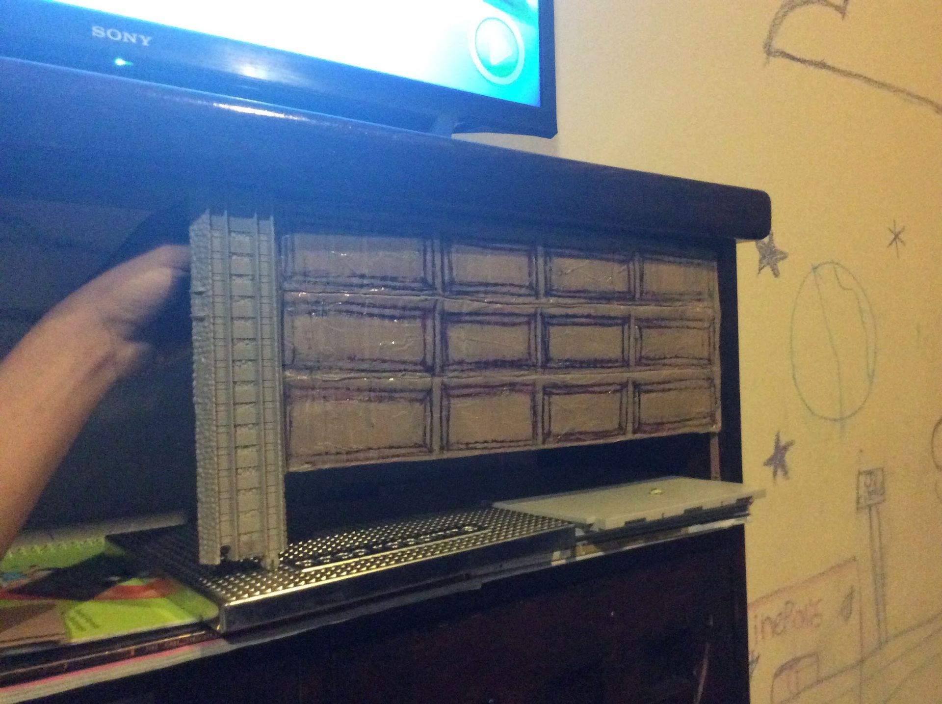 Person holding a gray building model next to a homemade cardboard wall on top of a dark cabinet.