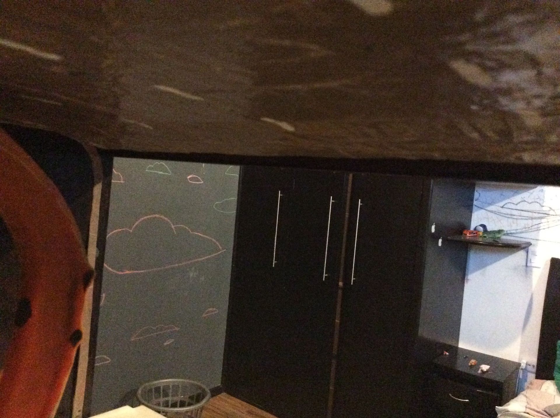 Interior view of a dark room with a closed black wardrobe, a wall with white cloud drawings, and an orange object.