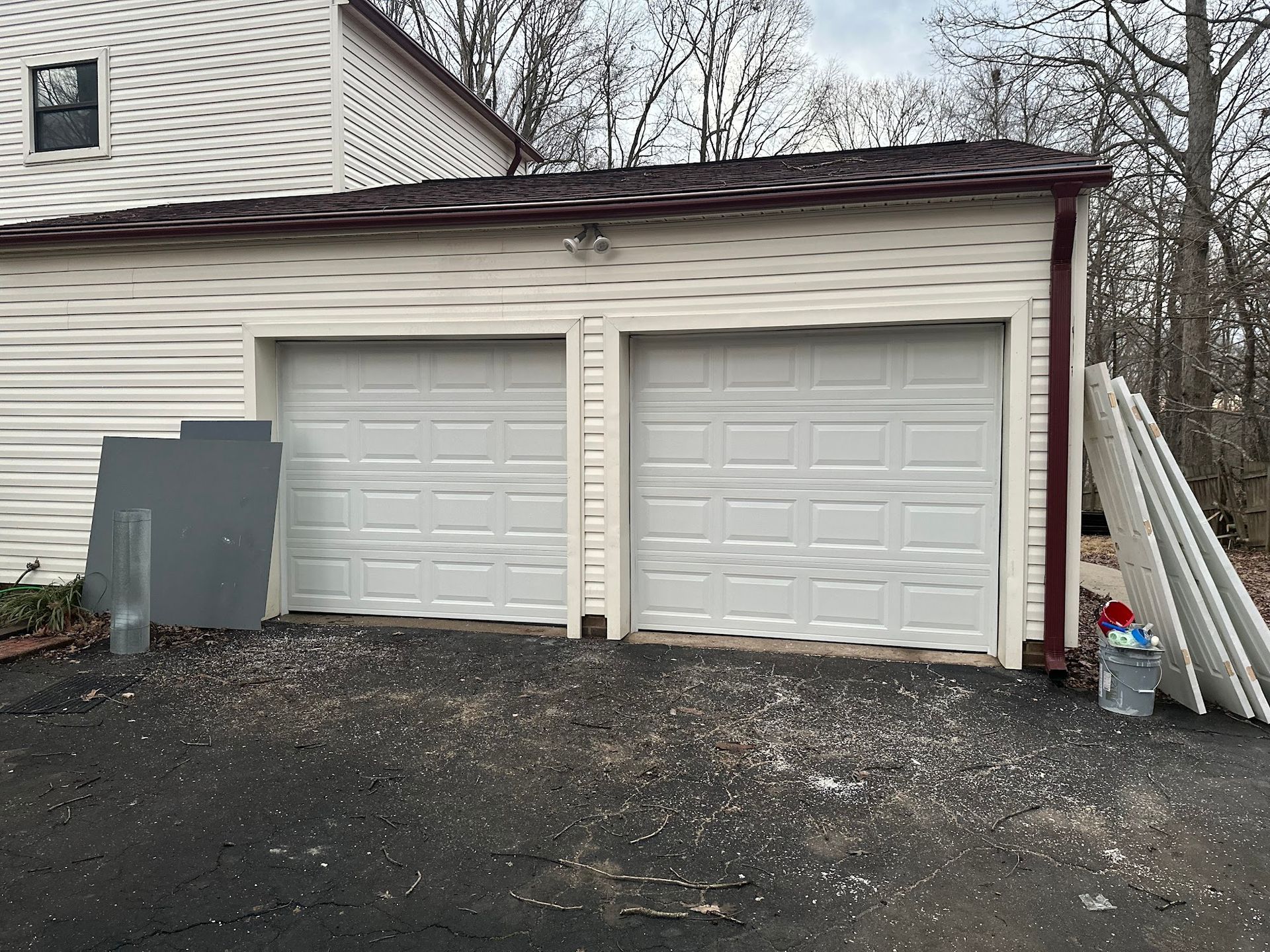 Two-car garage with white doors, surrounded by siding. Some materials and debris on the ground.