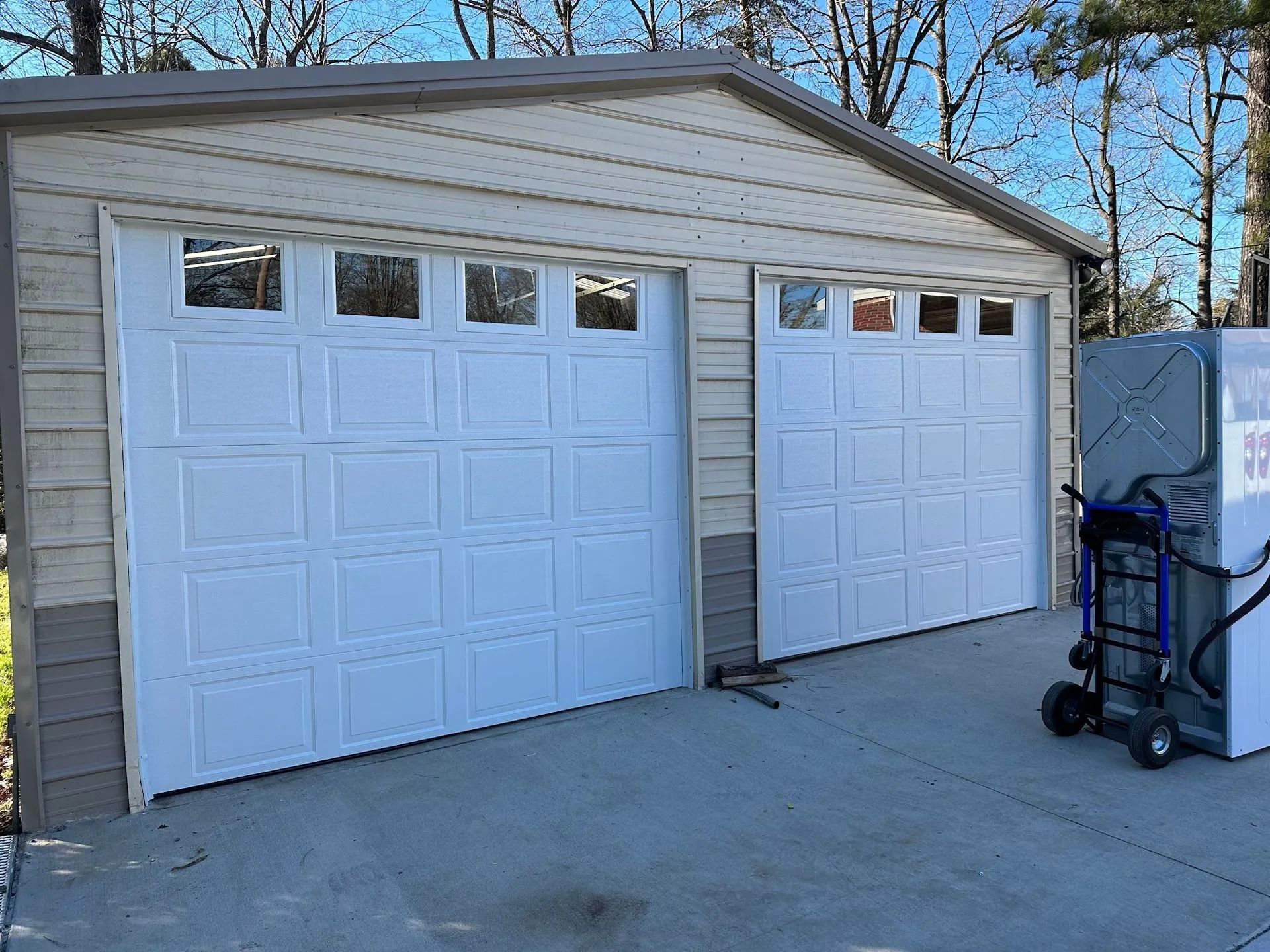 Two white garage doors on a beige house with a concrete driveway and a hand truck nearby