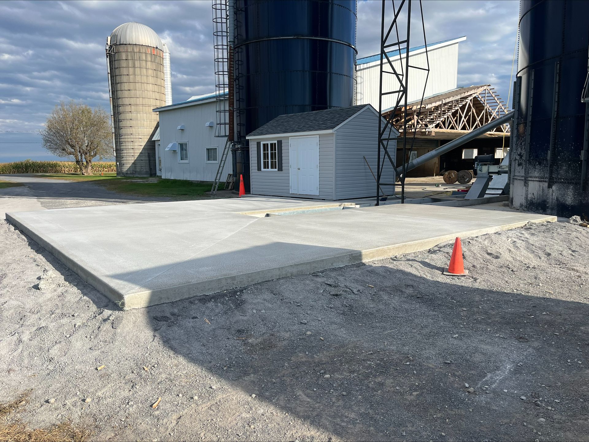 Une plateforme en béton est en cours de construction devant une ferme avec des silos et un hangar.