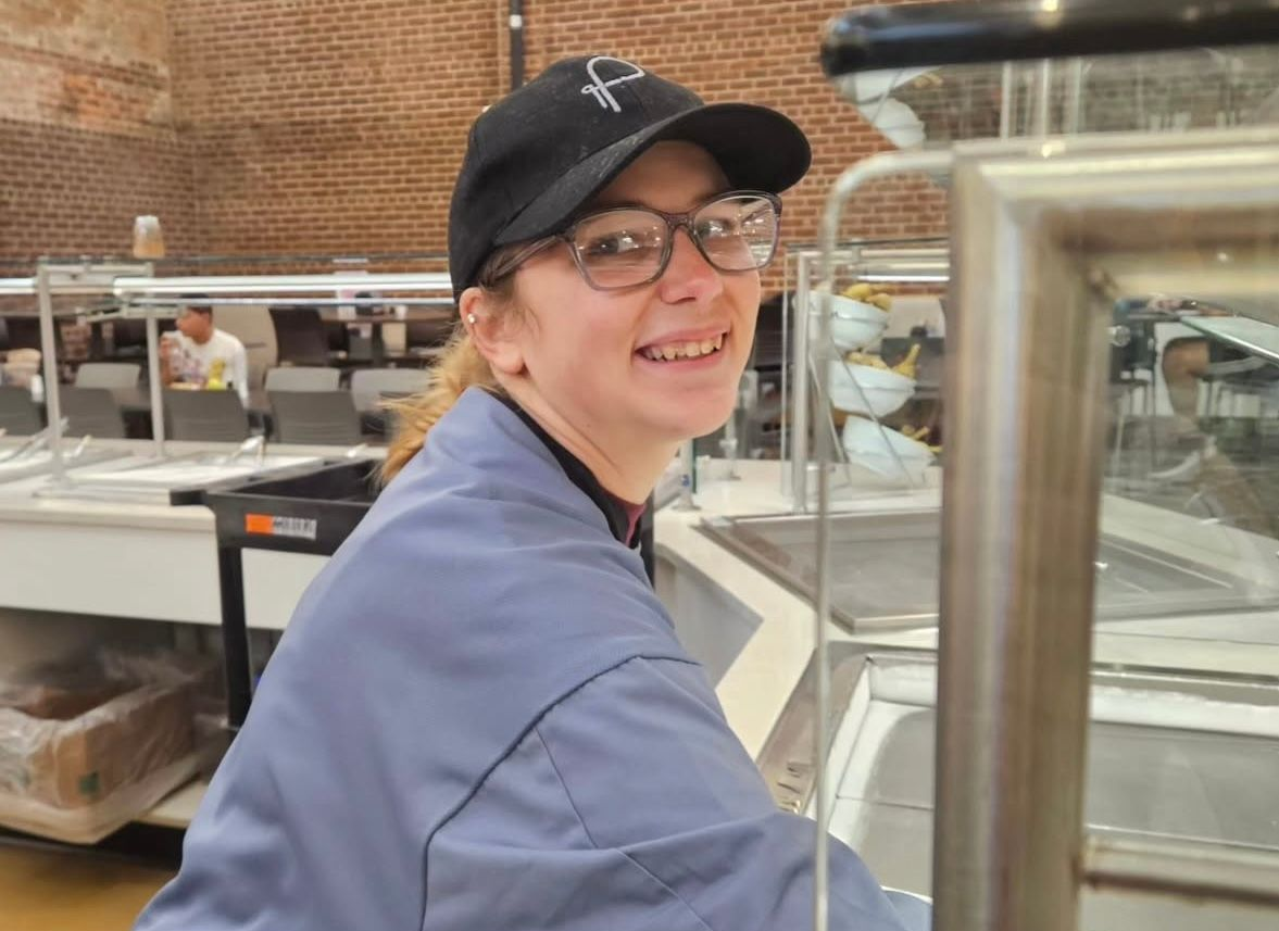 Woman in a blue uniform and cap smiles while serving food at a cafeteria.
