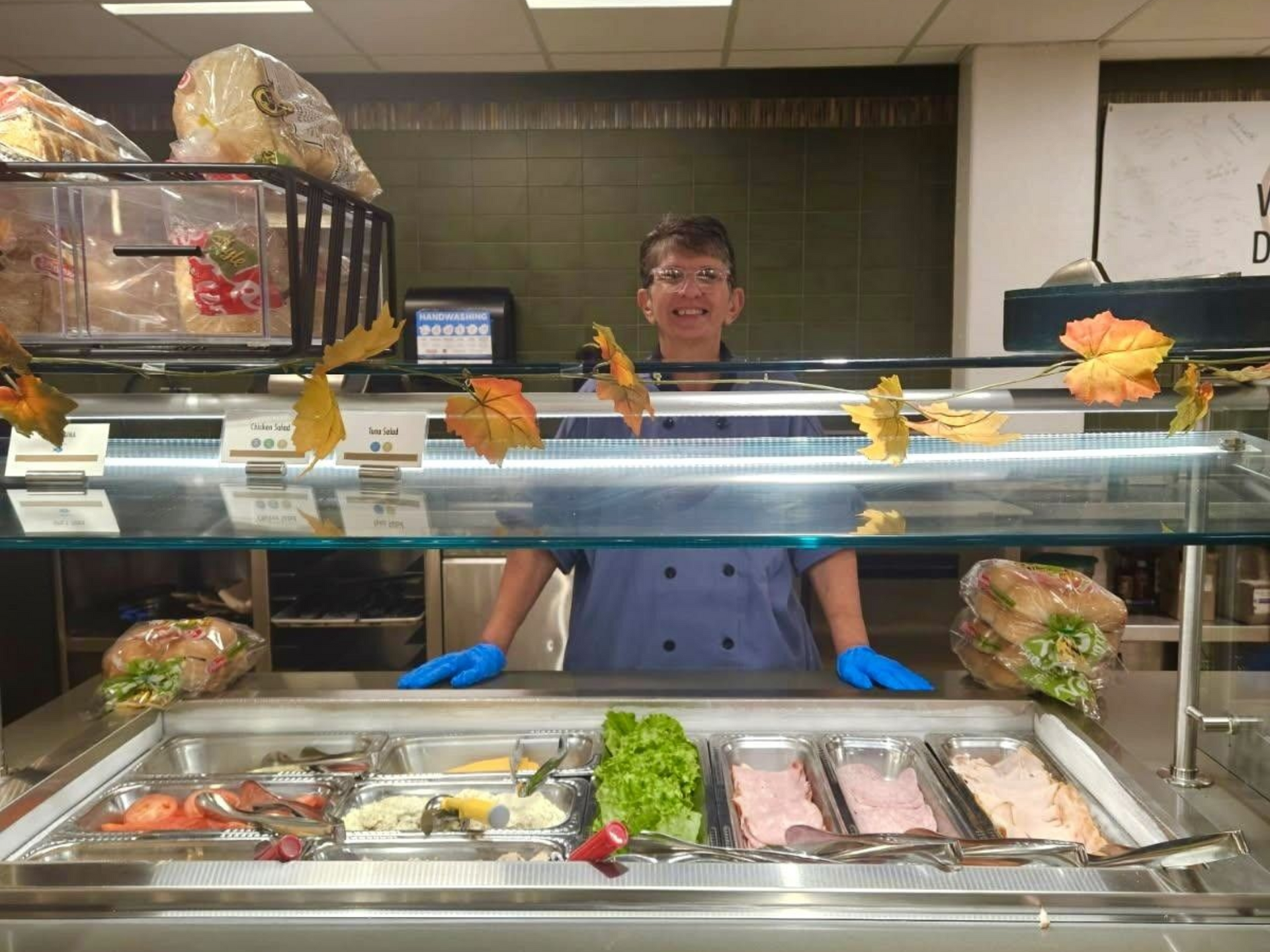 Person smiling behind a food display case with sandwiches, salads, and meats. They are wearing blue gloves.