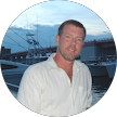 A man in a white shirt is standing in front of boats in a harbor.