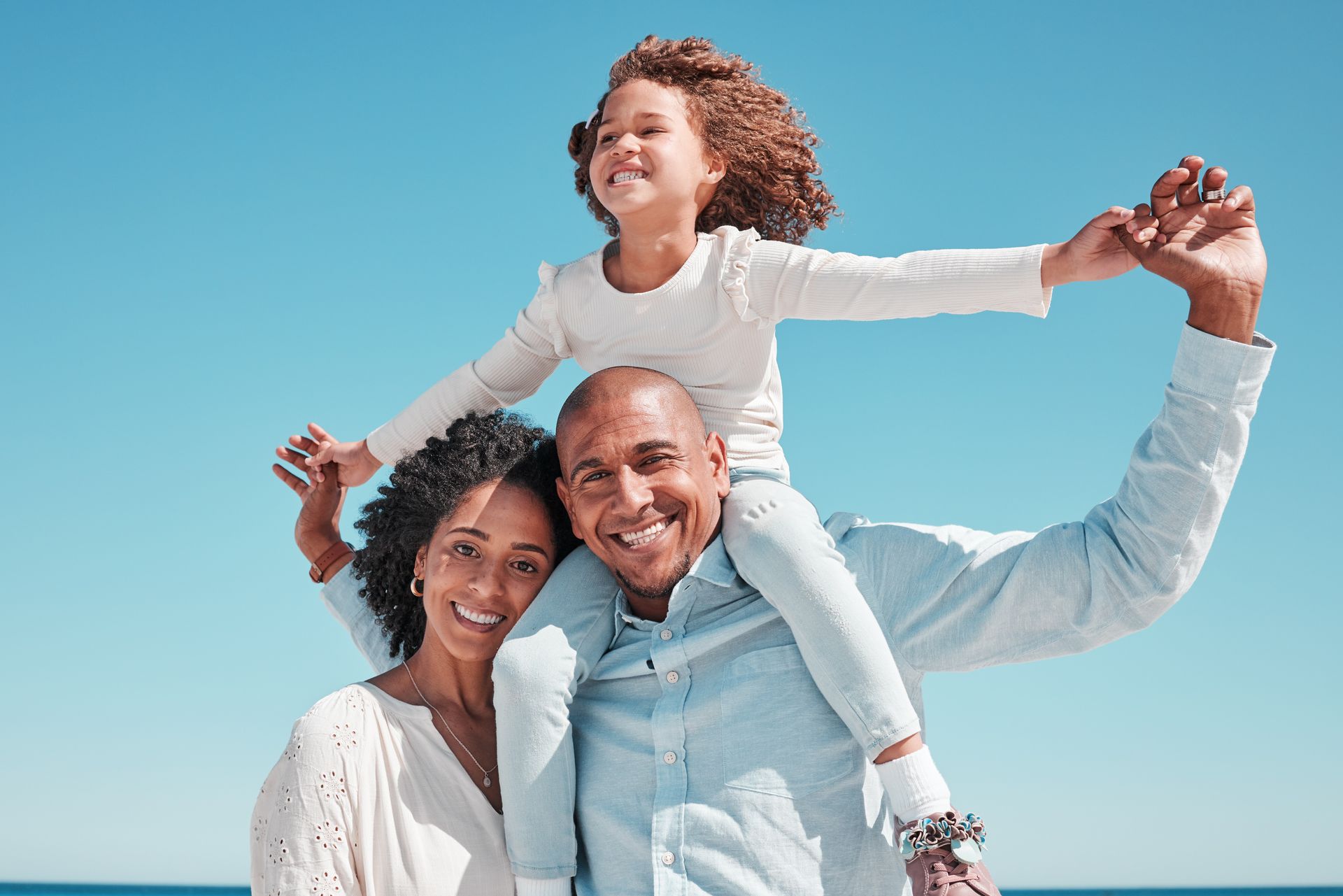 Happy family of three at the beach: father holding daughter on shoulders, mother smiling nearby, all against a blue sky.