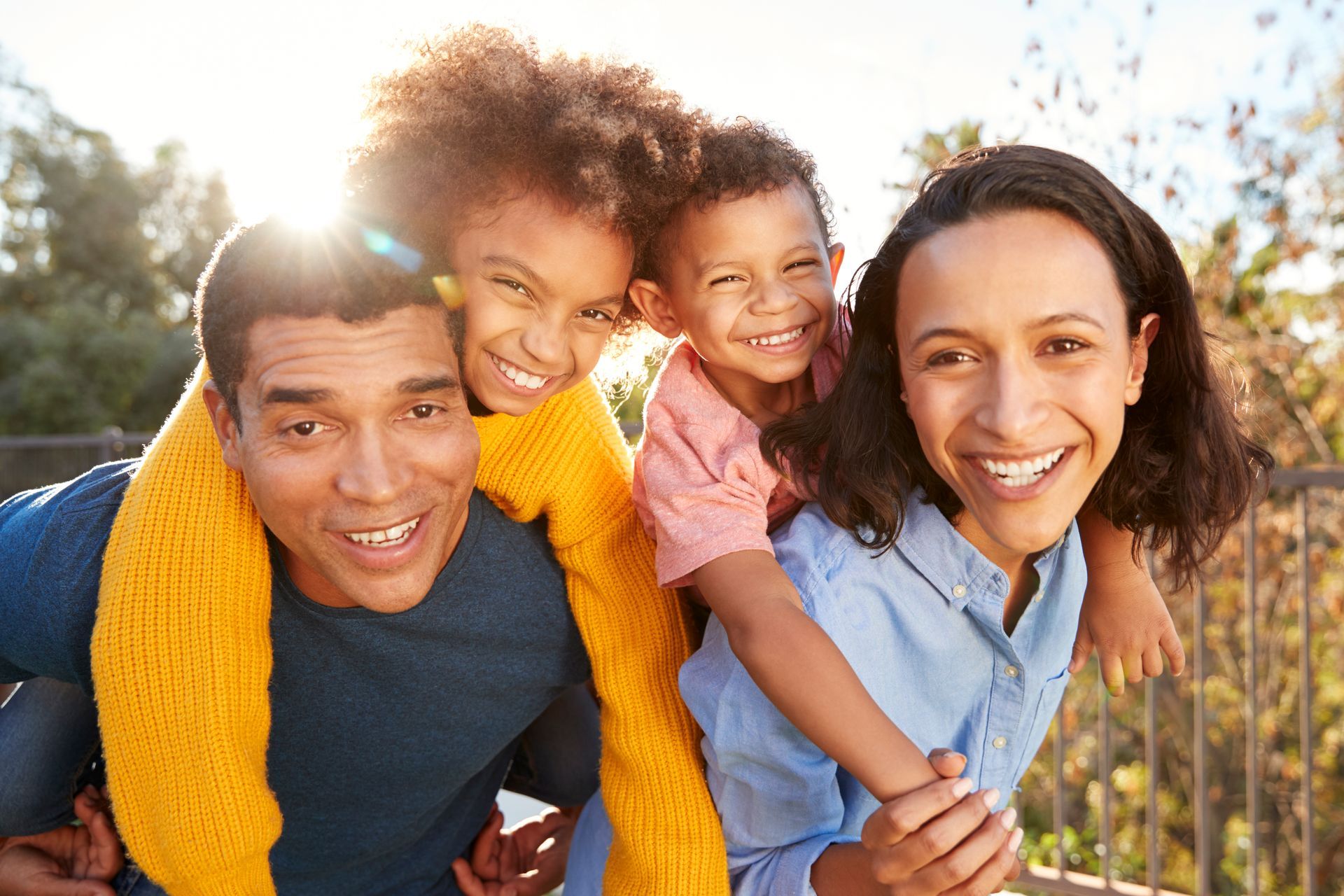 A smiling family of four outdoors; the parents giving piggyback rides to their two children.