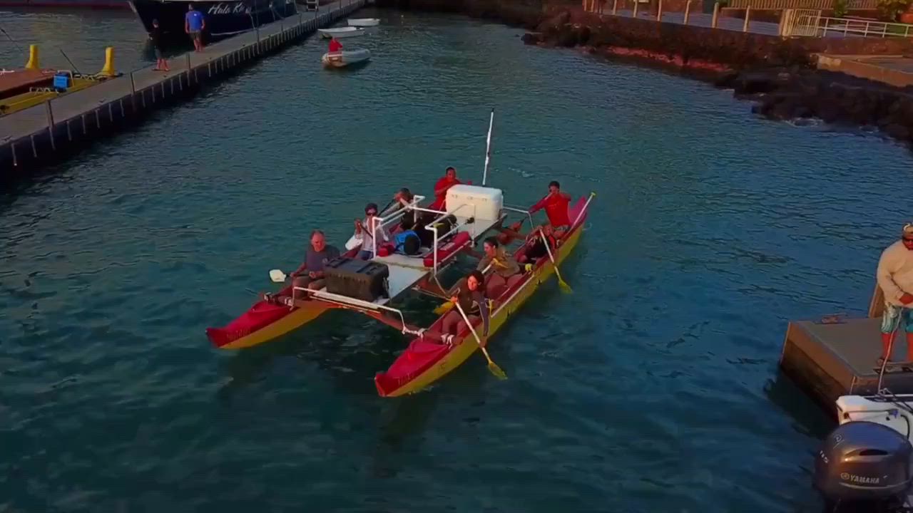 A red and yellow catamaran with people paddling in a harbor, near a pier.