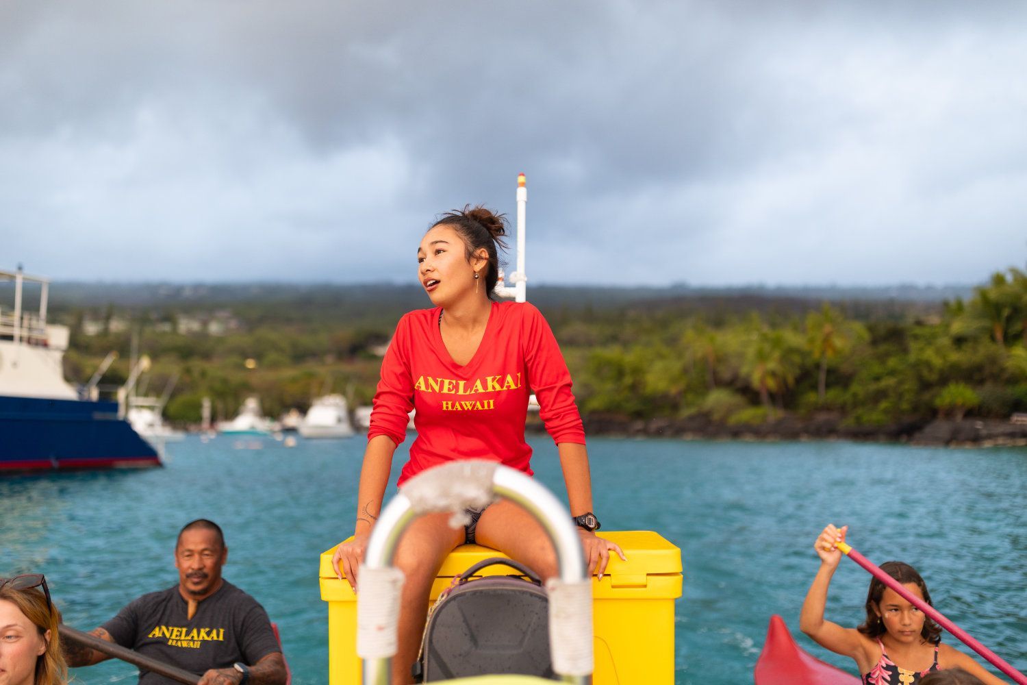 Woman on yellow buoy, singing on water, wearing red shirt, with rowers in background.