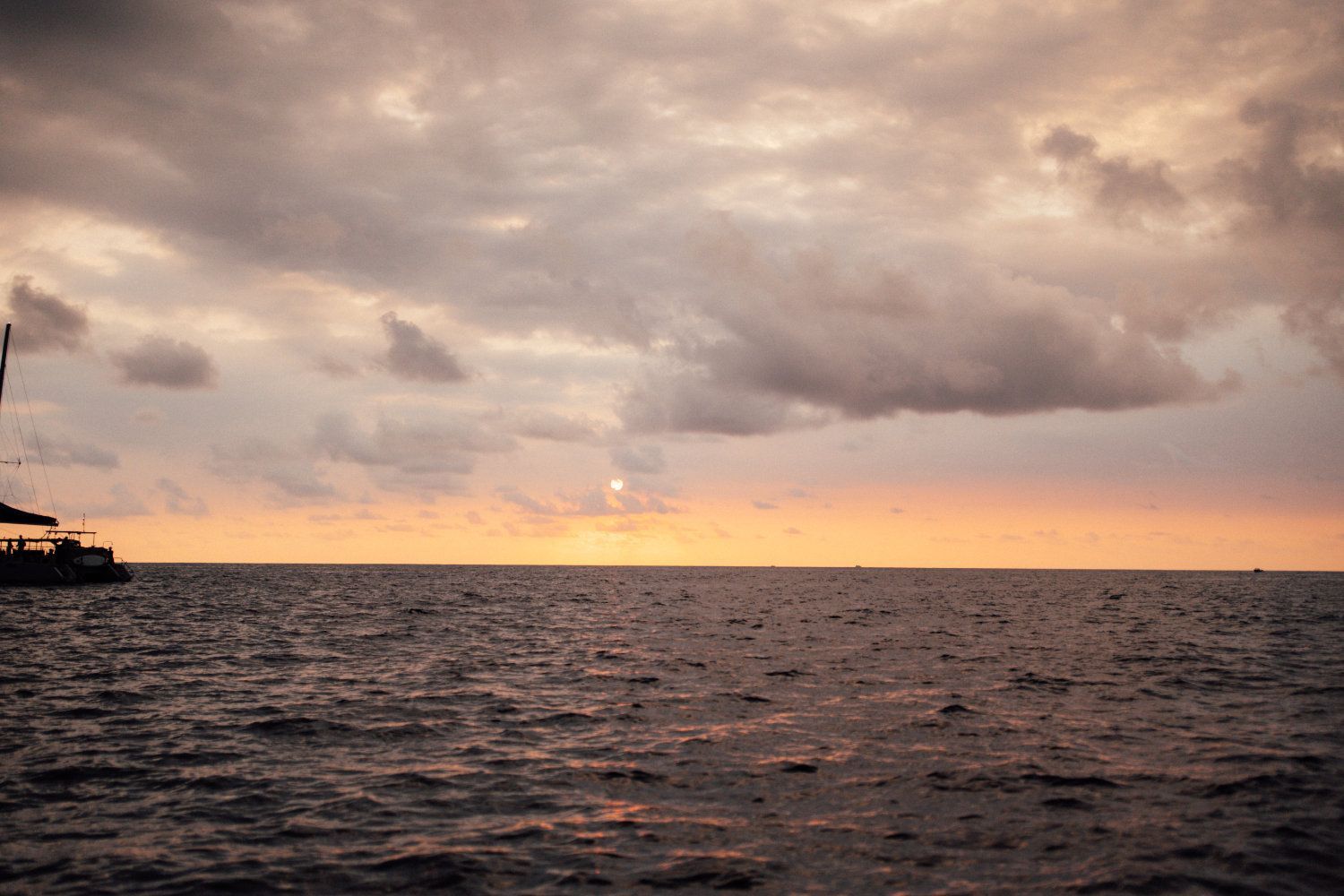 Ocean at sunset, cloudy sky; soft orange and grey hues. Boat silhouette on left.