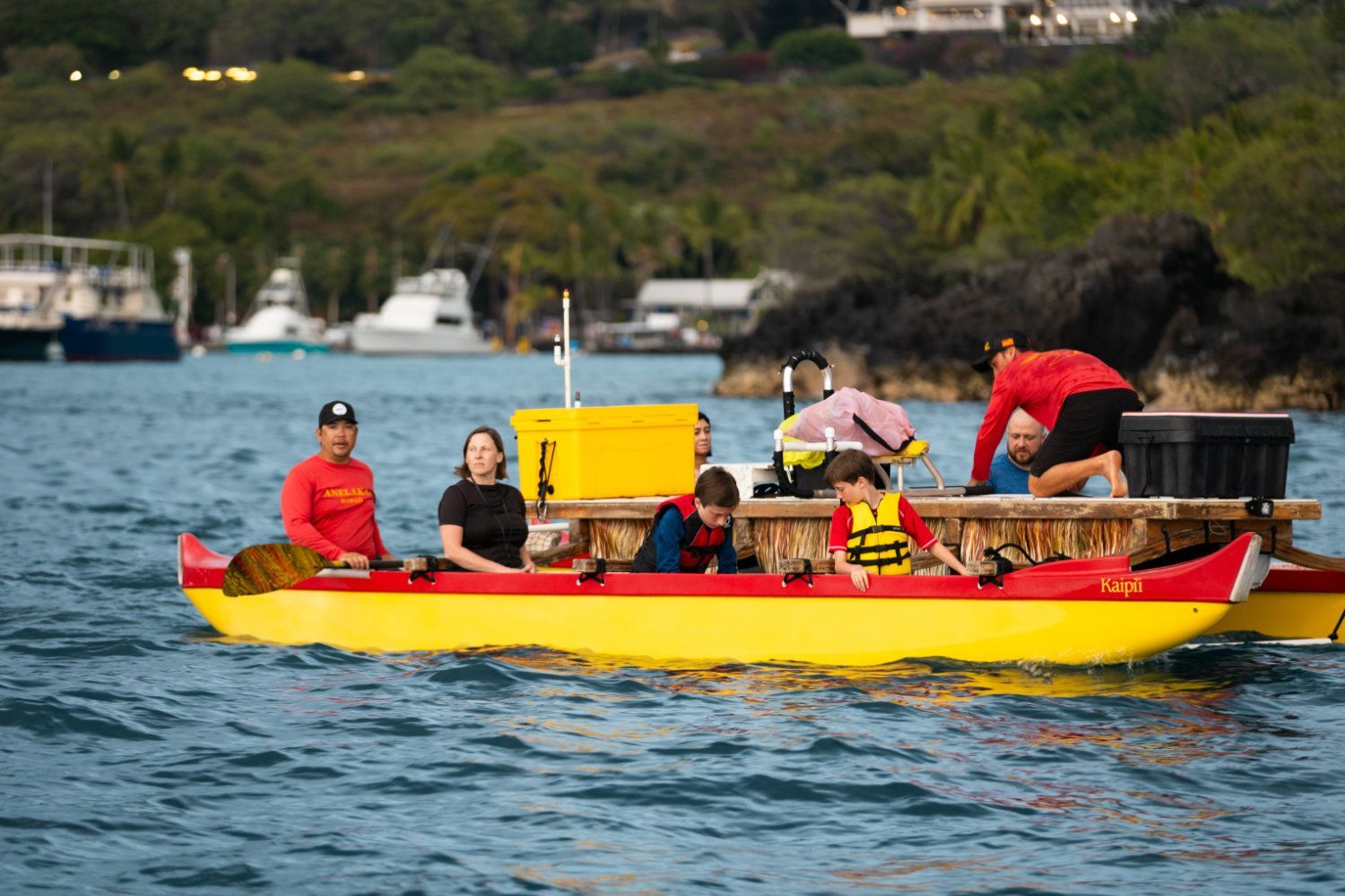 Yellow and red outrigger canoe with people; ocean setting, dark green shoreline.