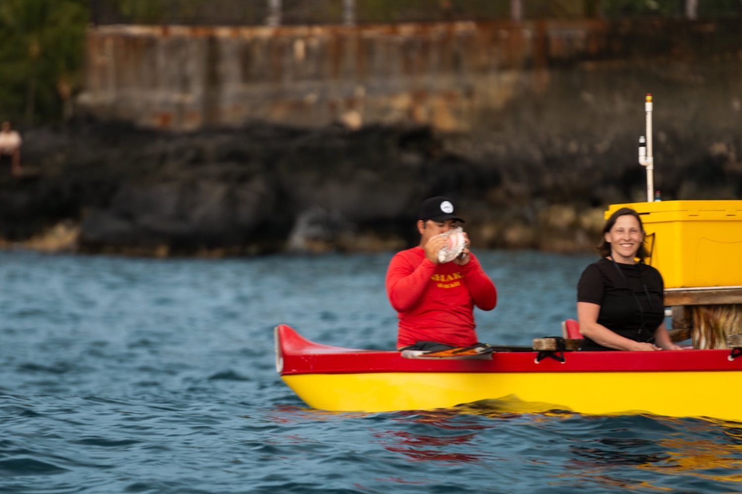 Man blowing conch shell, woman in boat on water. Red and yellow boat, dark rocks in background.