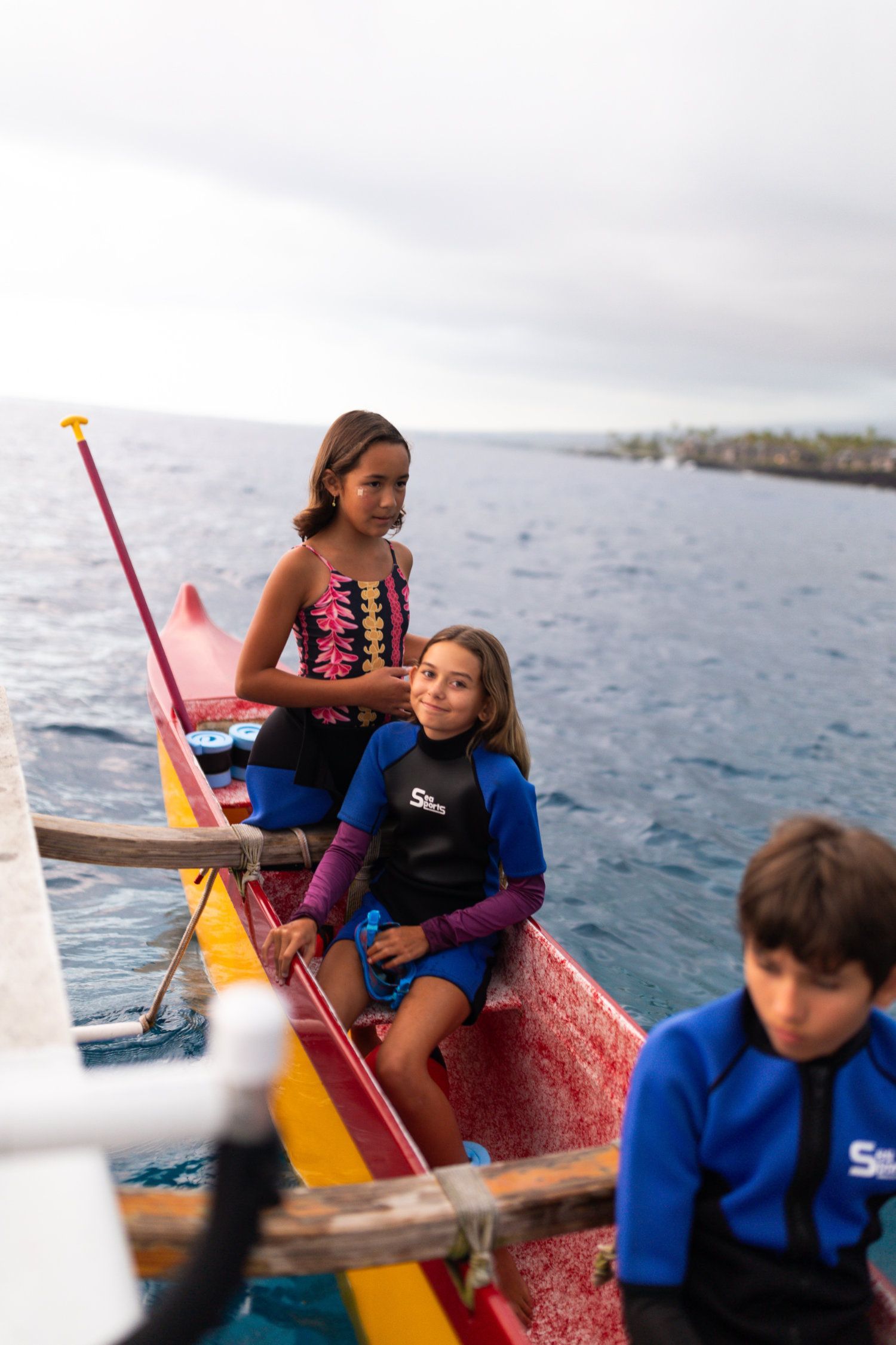 Children in boat on water; one in blue wetsuit smiles. Cloudy sky, ocean view.