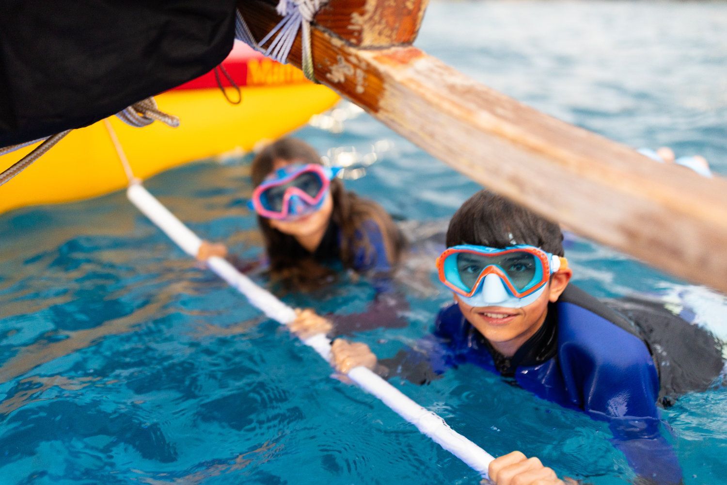 Two people in snorkel masks holding onto a white pole in blue water near a boat; smiling.