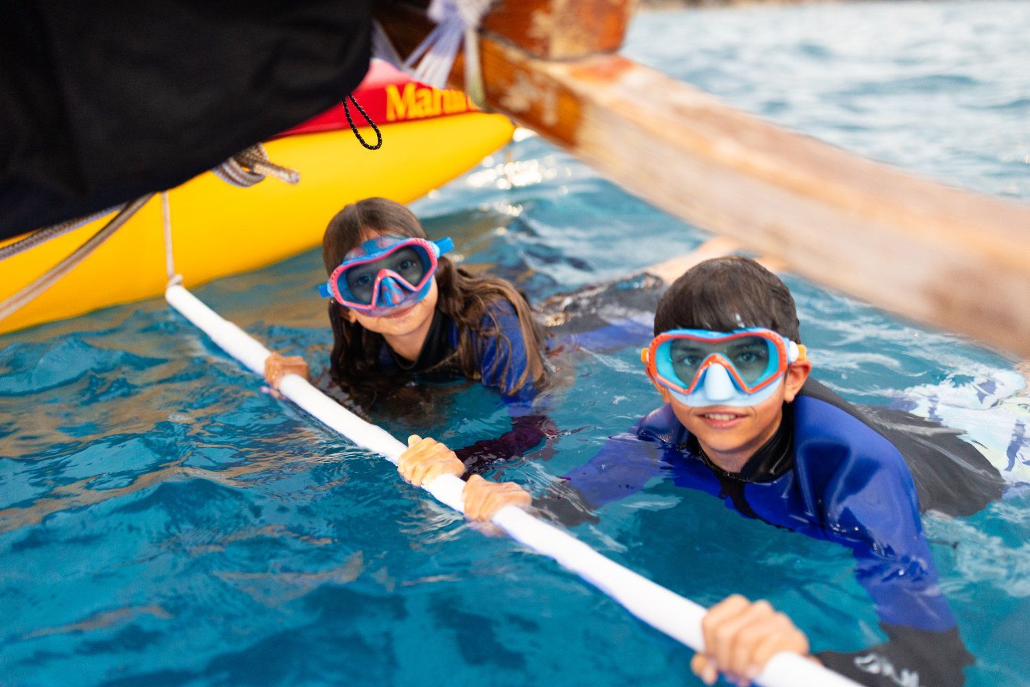 Two children in snorkeling gear in water, holding a white pole near a yellow boat.