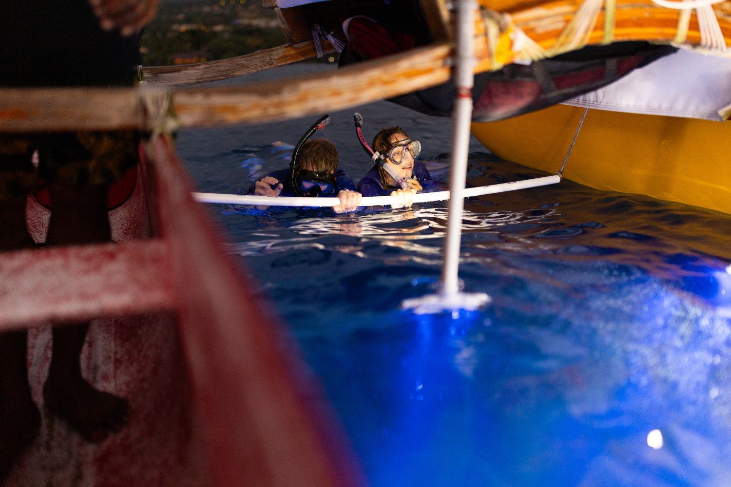 Two people snorkeling in blue water under a boat, holding a white rod.