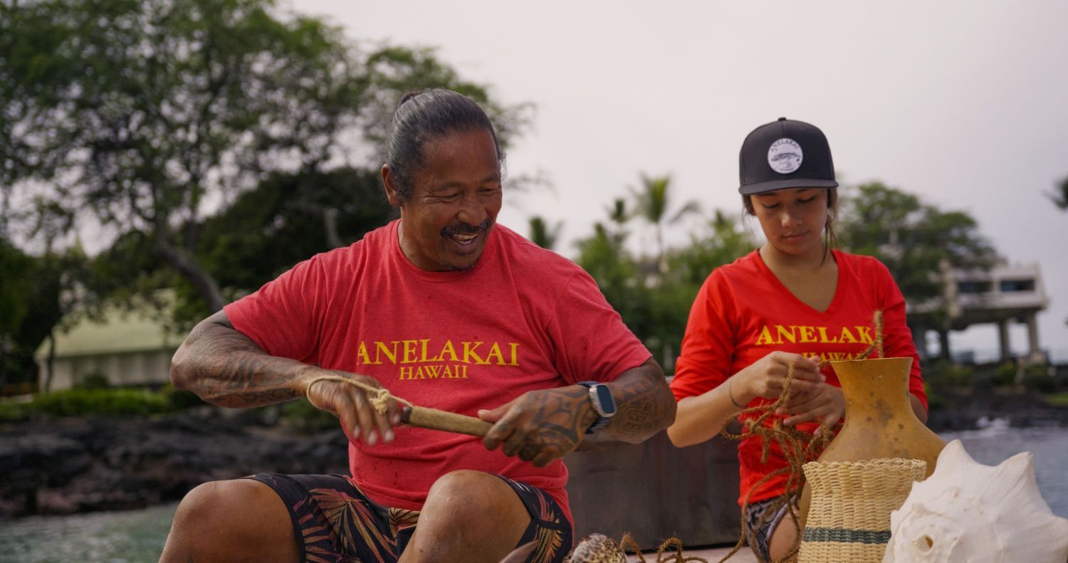 Two people in red shirts by the shore, crafting. One holds a wooden stick, the other works with string, near a container.