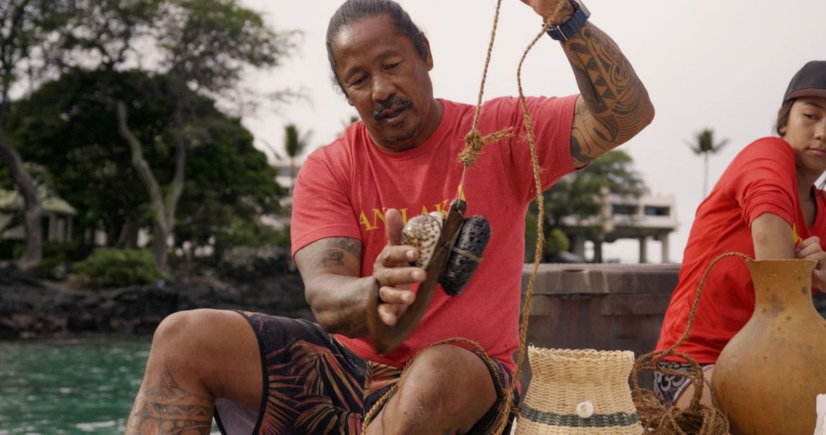 Man in red shirt holding objects on a dock. He's making knots. Other person nearby.