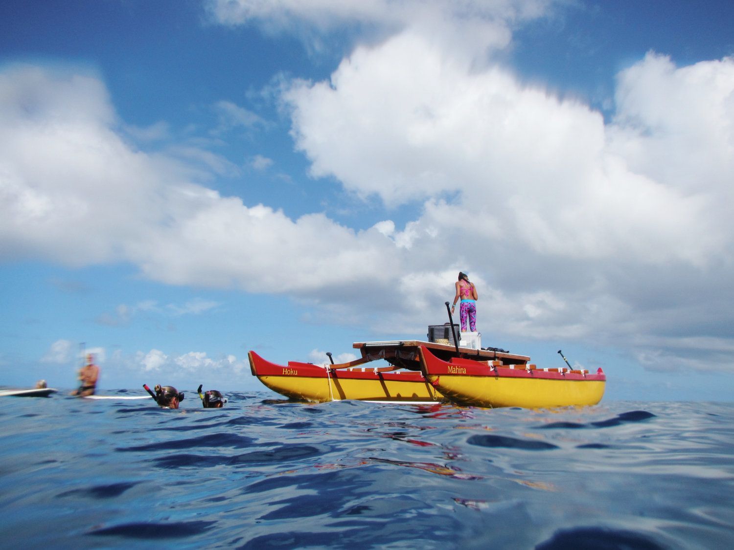 Yellow and red outrigger canoe on ocean under blue sky with several people.