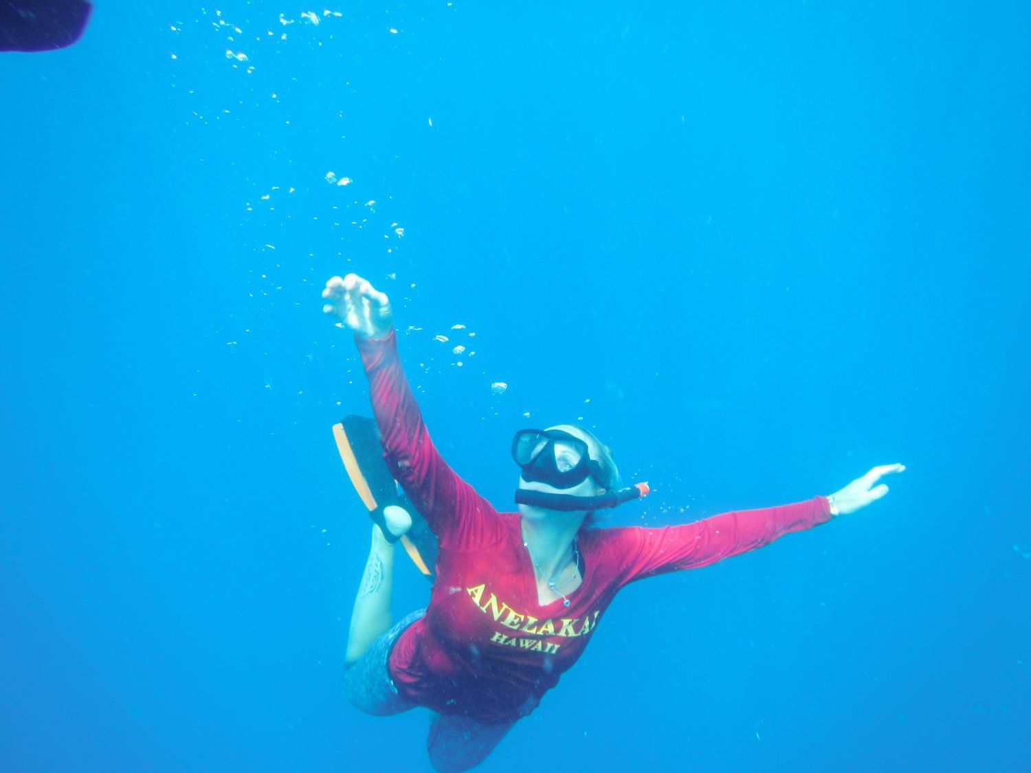Person snorkeling underwater, arms outstretched, wearing red shirt and fins, surrounded by blue water.