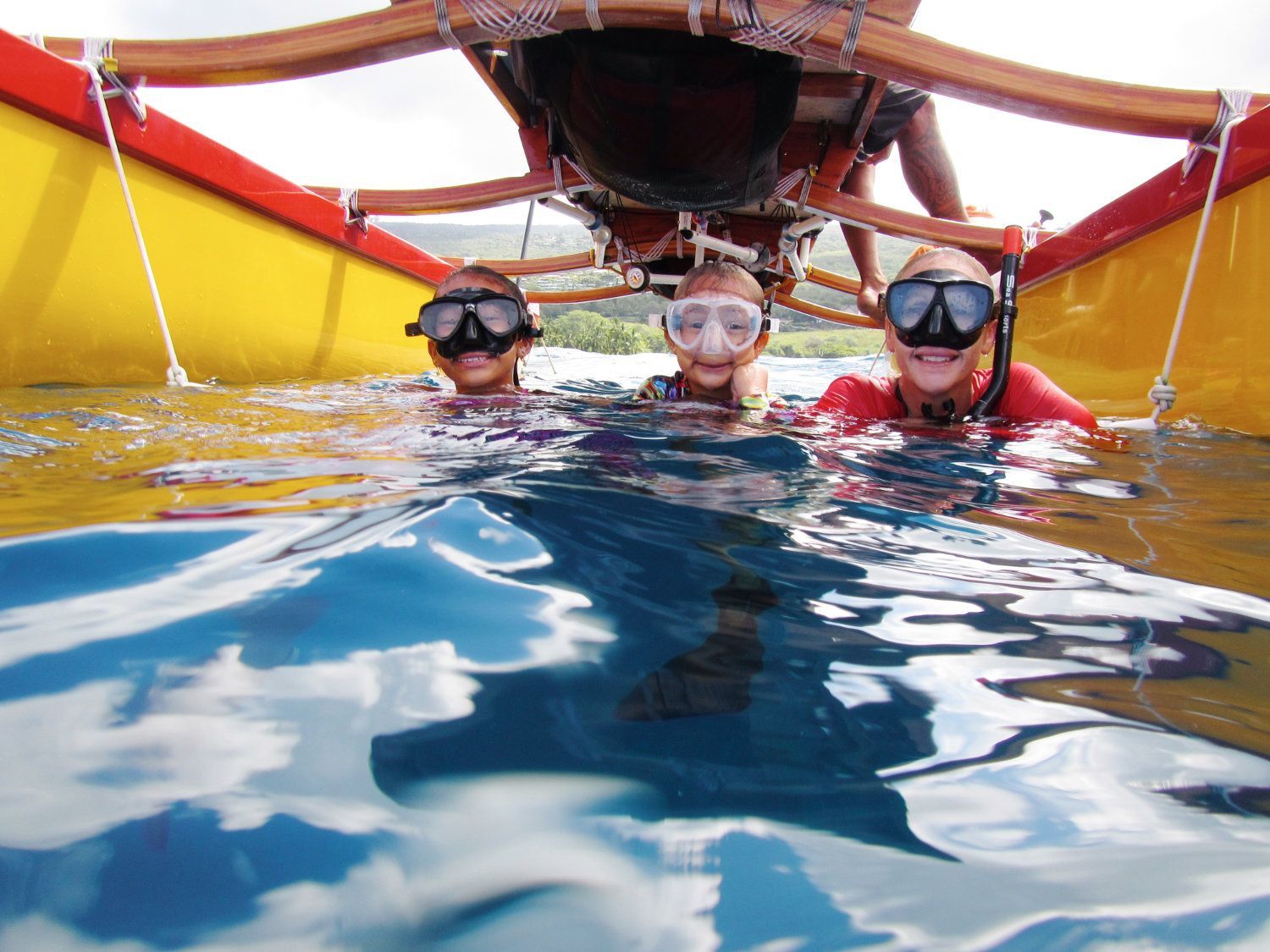Three people snorkeling under a boat, smiles and goggles visible above water.