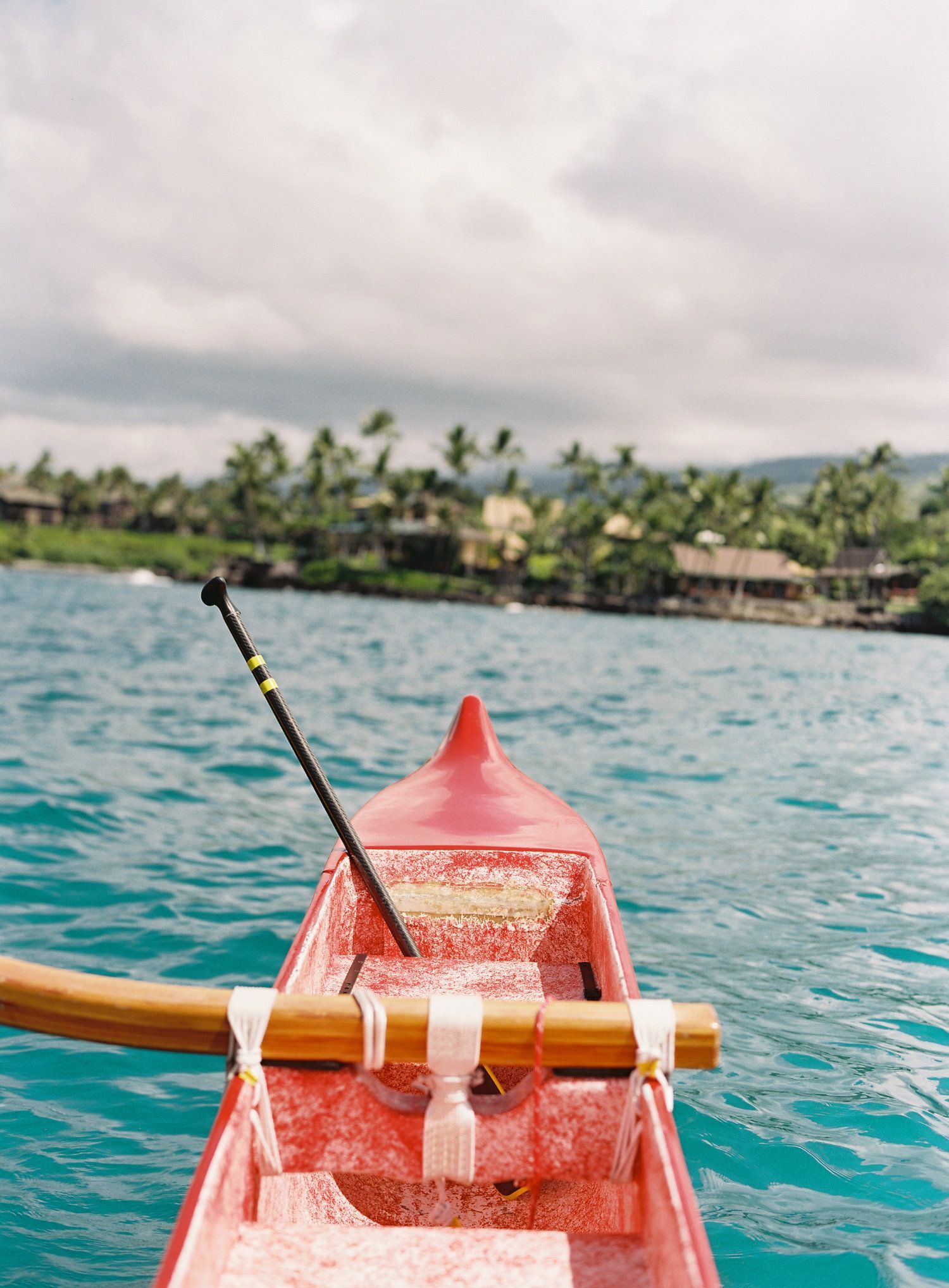 Red outrigger canoe on turquoise water, paddle extending. Lush green shoreline with buildings in background.