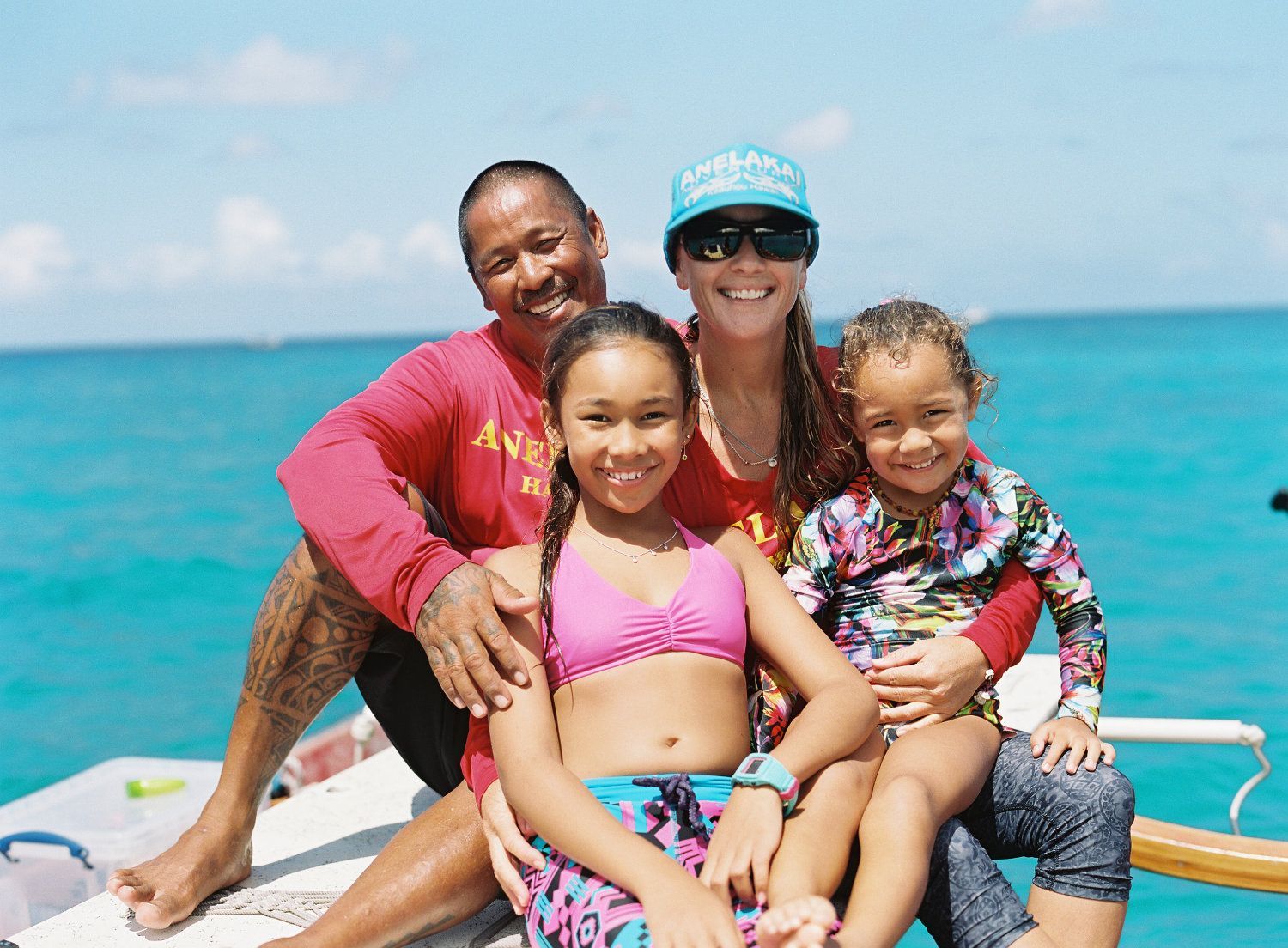 Family smiles in the sun on a boat. Ocean water in background. Two children sit with parents.