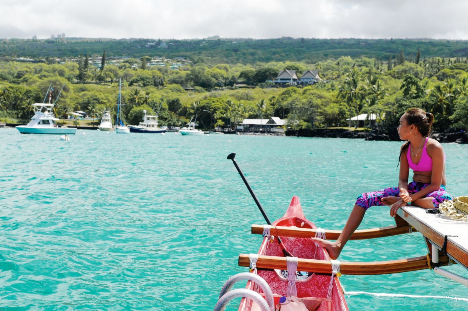 Young person on outrigger canoe, turquoise water, boats, lush green shoreline.
