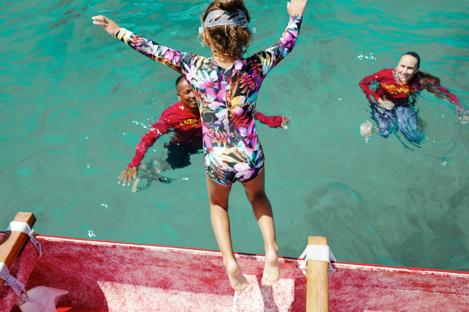 Child jumping into turquoise water from a red boat, arms outstretched. Two people swim nearby.