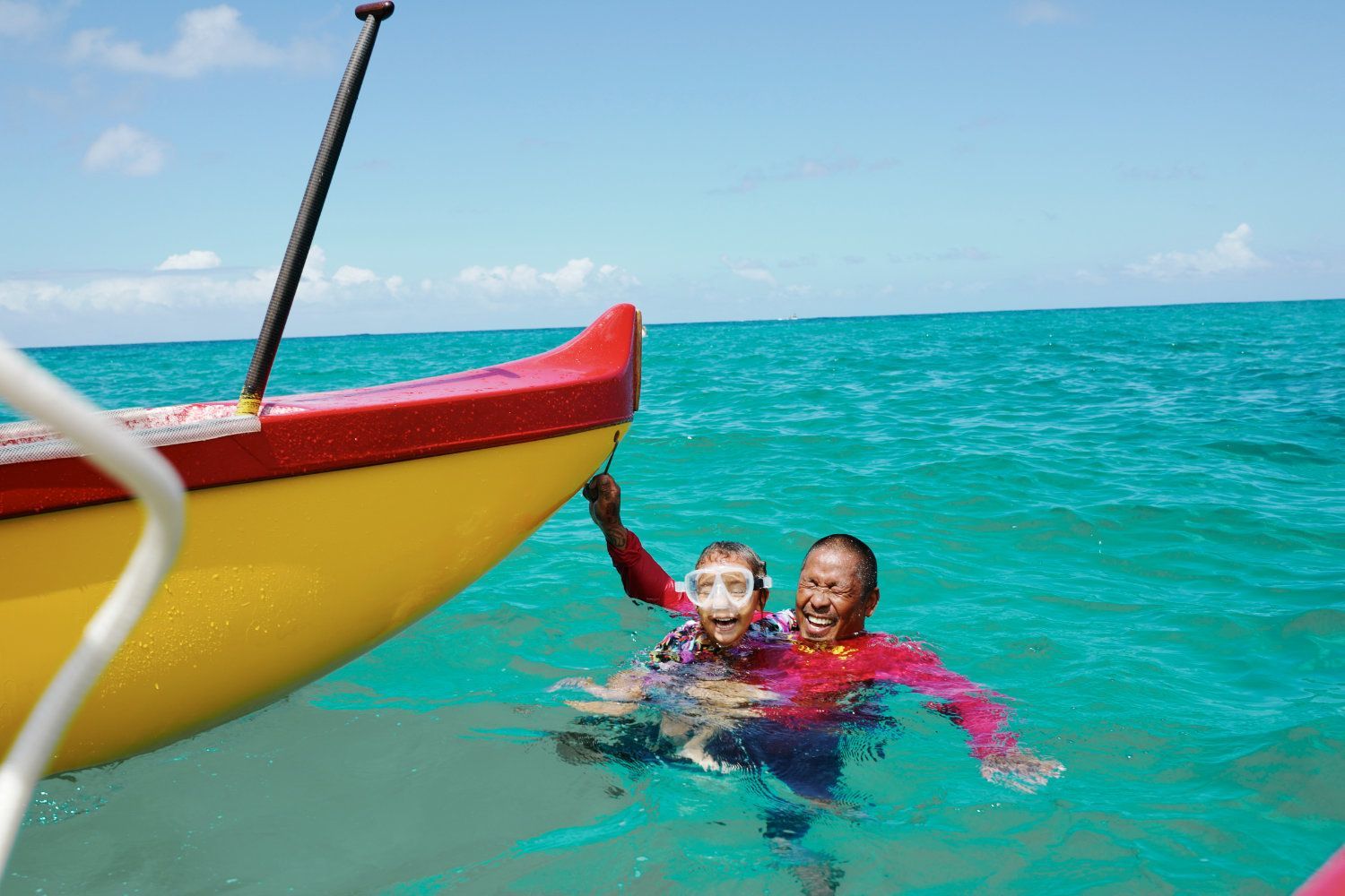 Two people in turquoise water near a yellow and red boat. One person is wearing a swim mask and smiling.