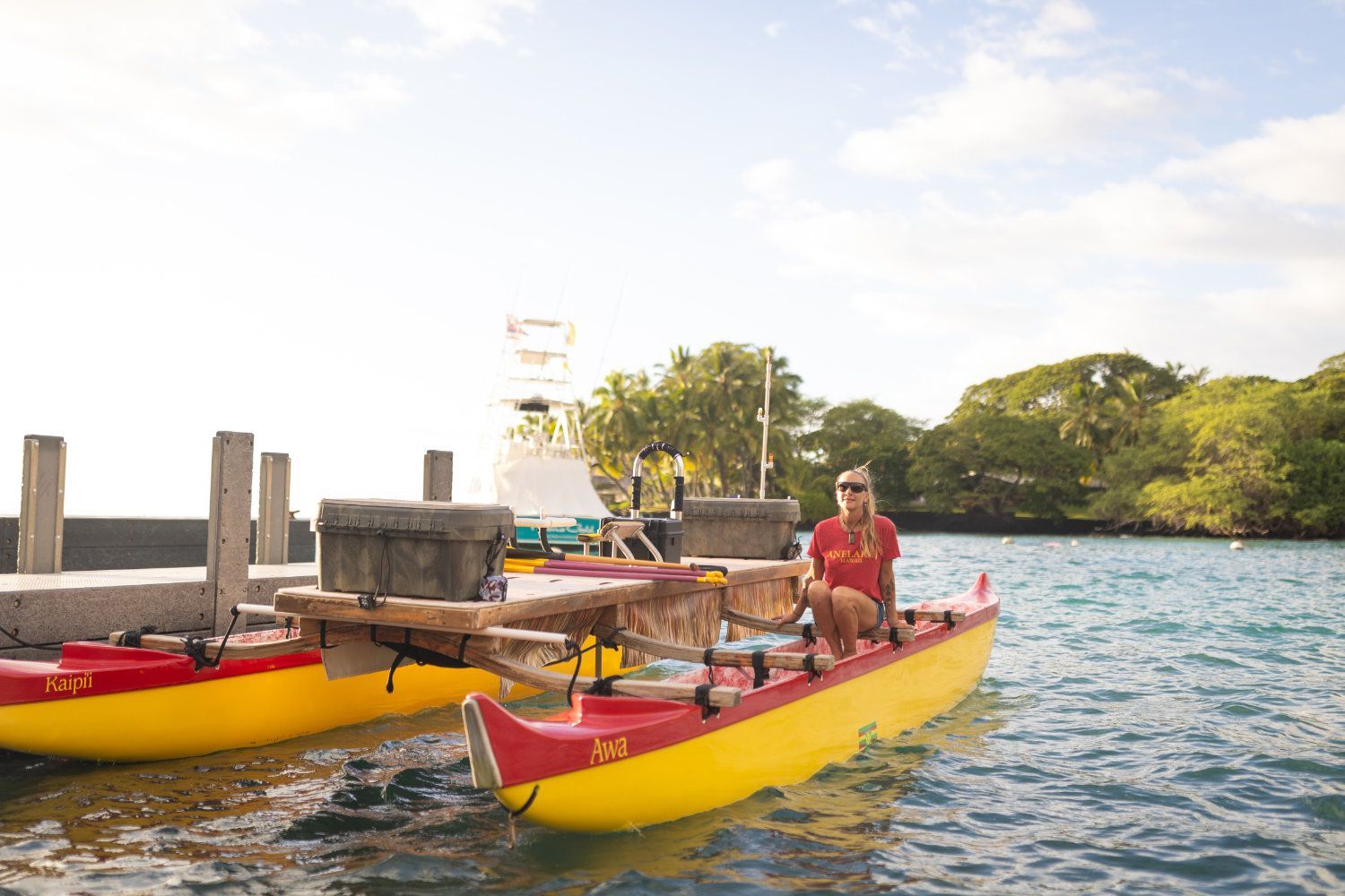 Woman in red shirt on a yellow and red outrigger canoe at a dock on sunny day.