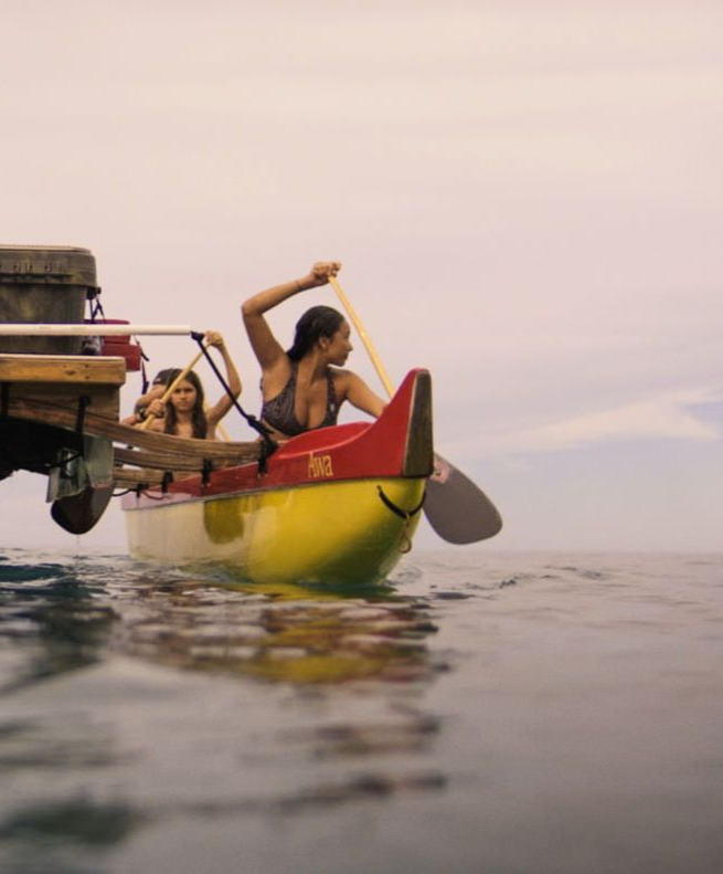 People paddling a yellow canoe on water. A woman paddles with a raised arm; two others sit in the boat.