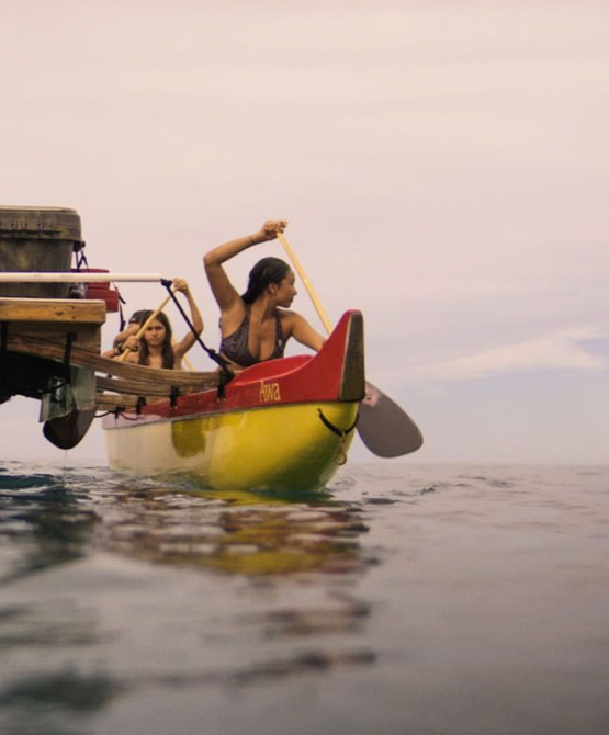 People paddling a yellow canoe on water. A woman paddles with a raised arm; two others sit in the boat.