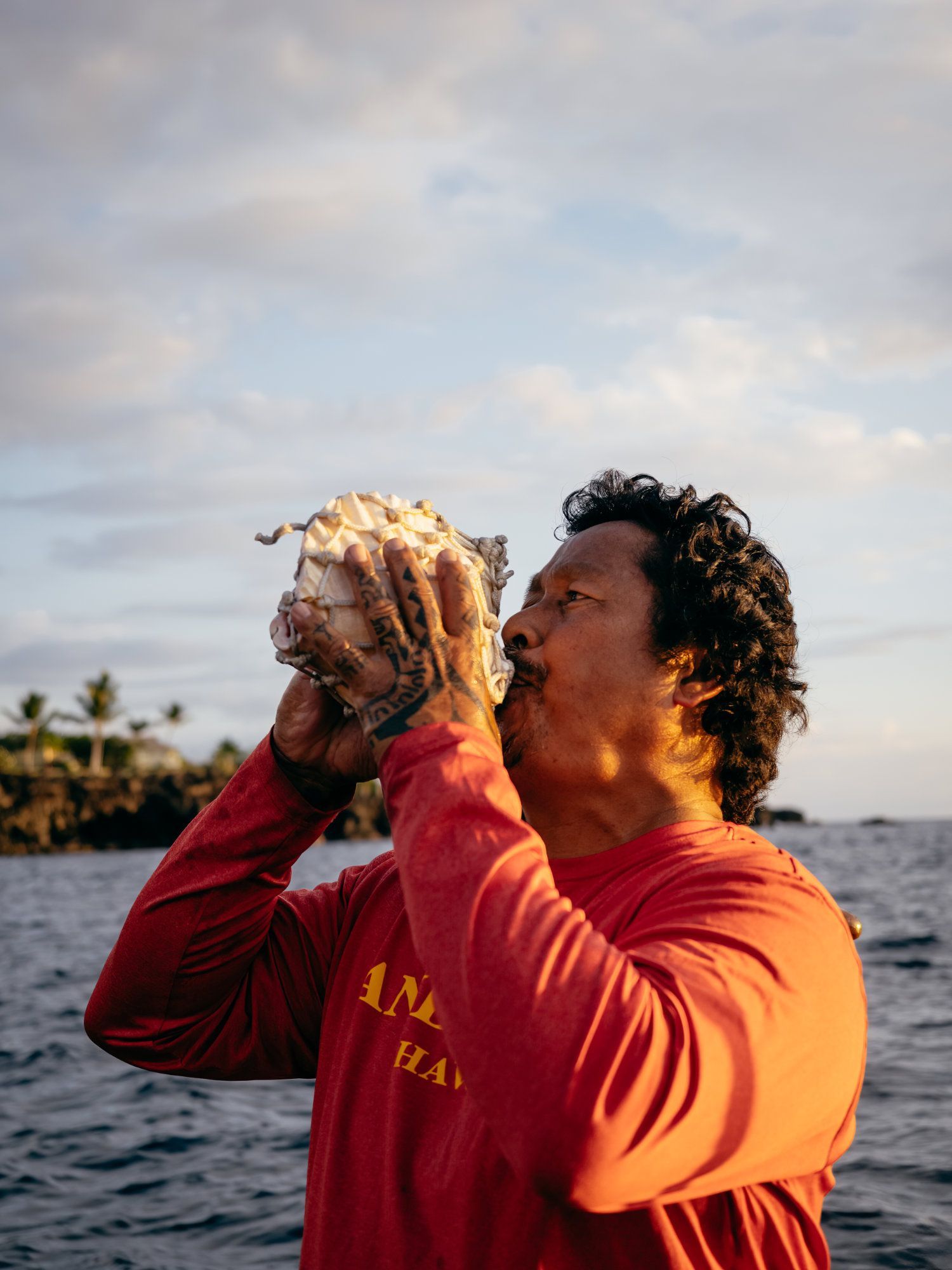 Man blowing into conch shell near water, wearing a red long-sleeve shirt.
