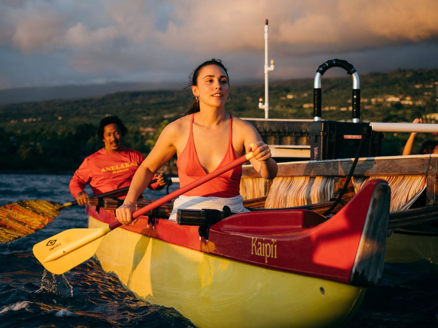 Woman paddling a yellow and red outrigger canoe. Another person in the background, near a dock.