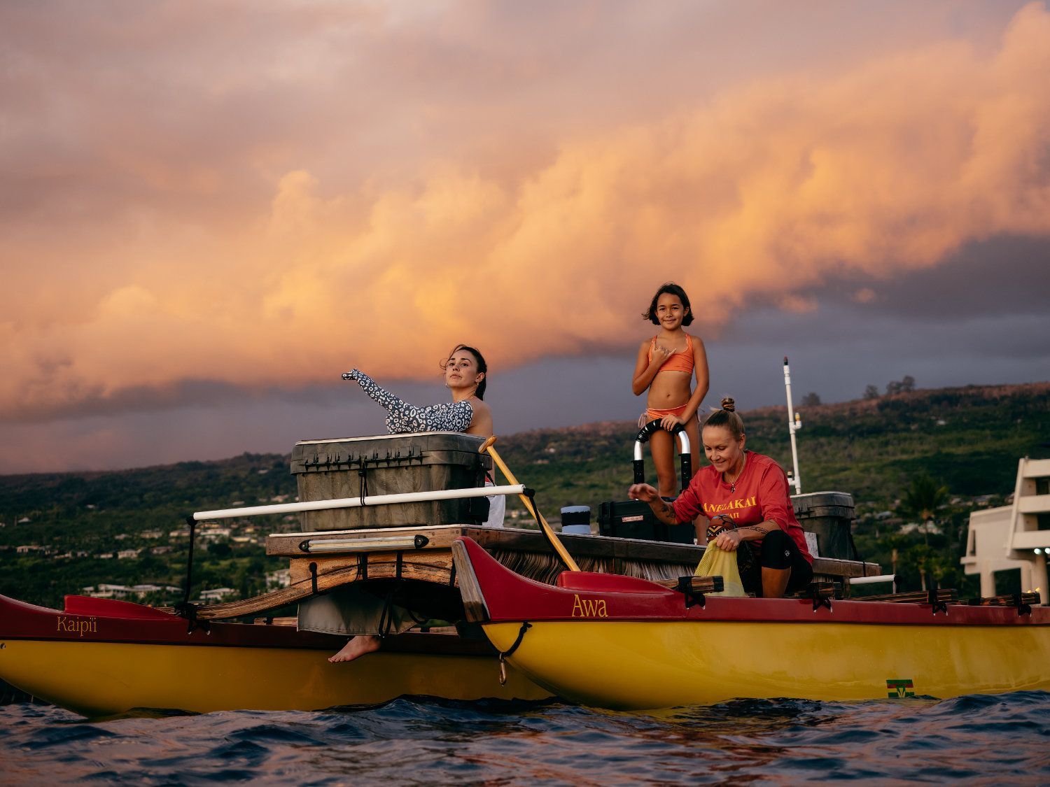 People on a yellow and red catamaran with a sunset backdrop. One raises arm, another stands, one rows.