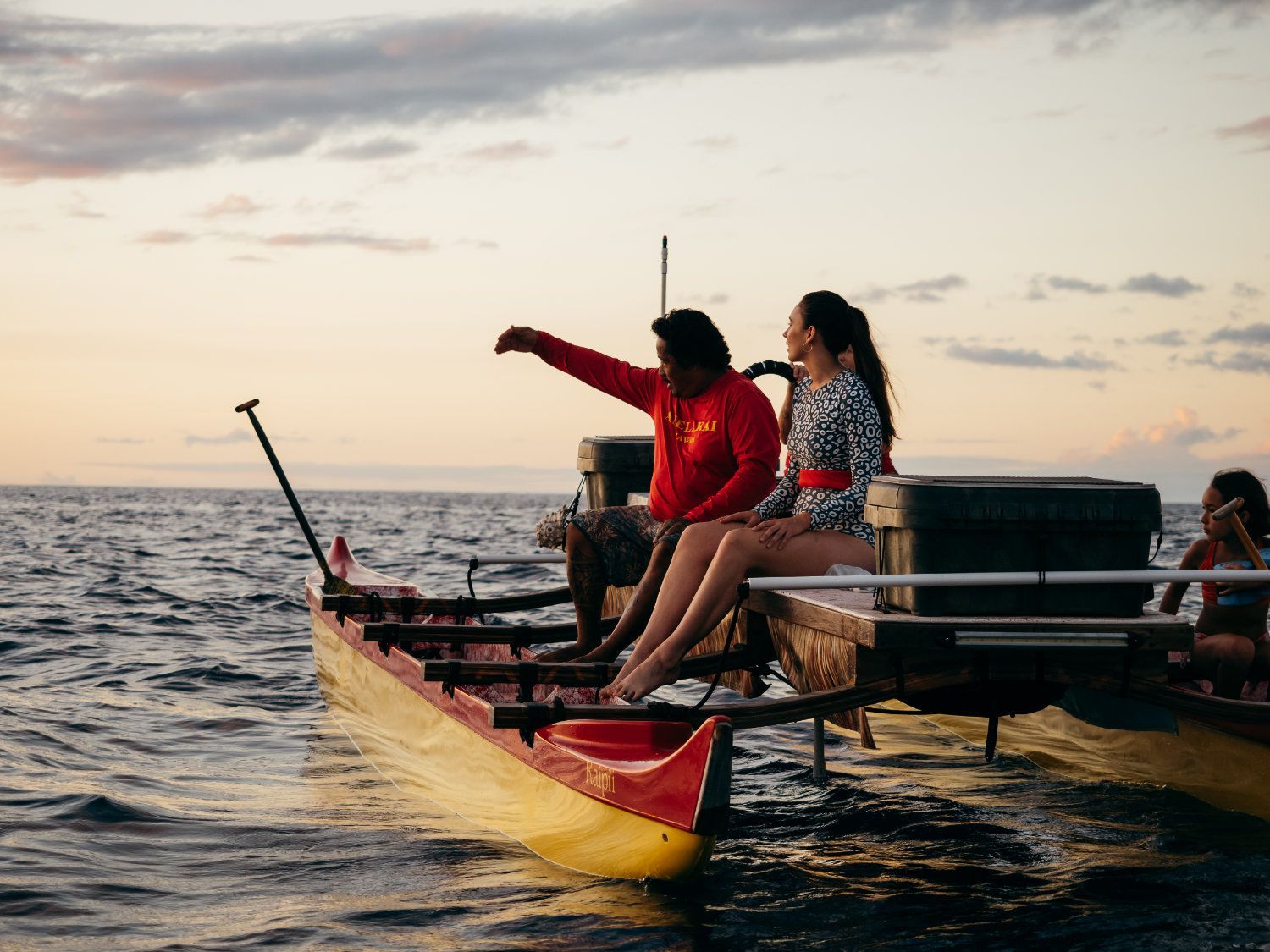 People on a yellow outrigger canoe at sea, one pointing, sunset colors.
